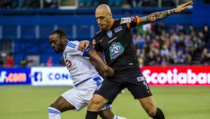 A Mountain view of Montreal City, Canada in the fall. The sky is clear and it is a sunny day. A cluster of gray, multiple-sized buildings are visible in the center of the photo. Lush, fall foliage lines the front of the image, and a body of water is visible in the back of the photo.